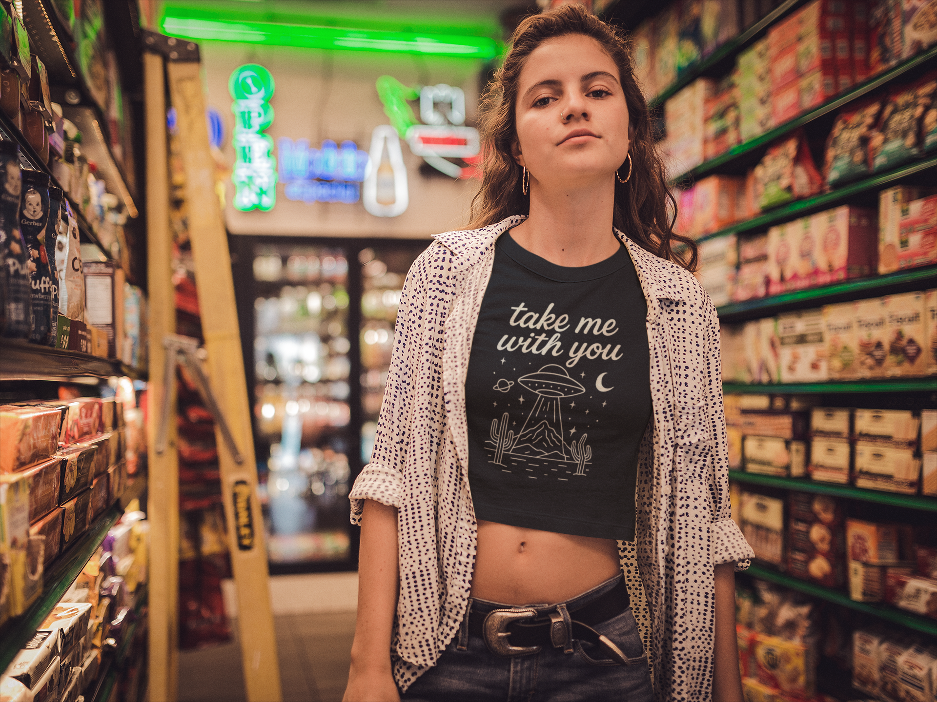 Woman standing in a neon-lit grocery aisle wearing Barely Functioning Co. black crop top with UFO design and text 'Take Me With You.' Alternative streetwear with surreal, cosmic vibe.