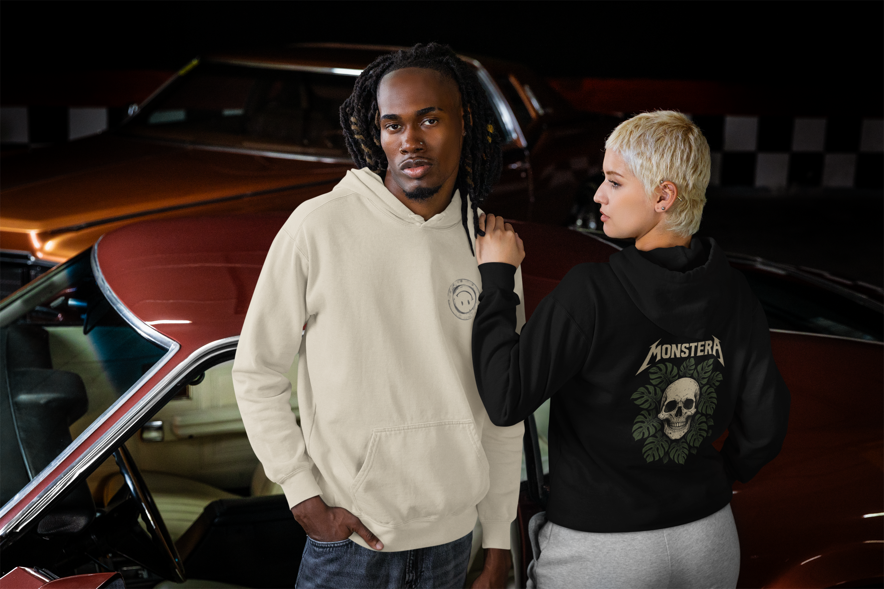 Man and woman wearing Barely Functioning Co. hoodies — beige hoodie with upside-down smiley logo on front, black hoodie with 'Monstera' skull and leaves design — edgy streetwear look in front of classic car.