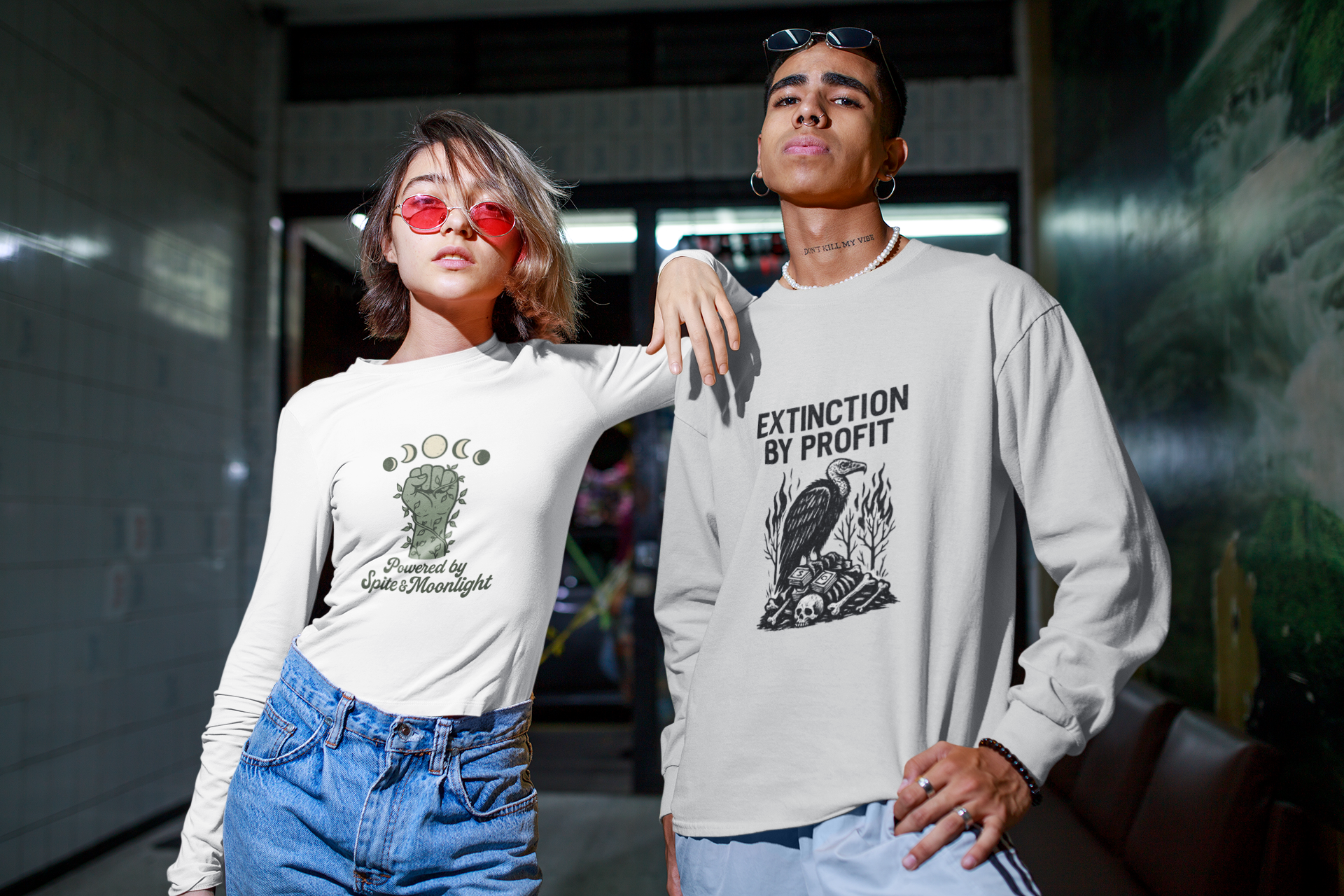 Two models wearing Barely Functioning Co. long sleeve shirts. Left model in white top with green graphic reading 'Powered by Spite & Moonlight.' Right model in white top with bold black design reading 'Extinction by Profit.' Alternative streetwear with sarcastic activist edge.
