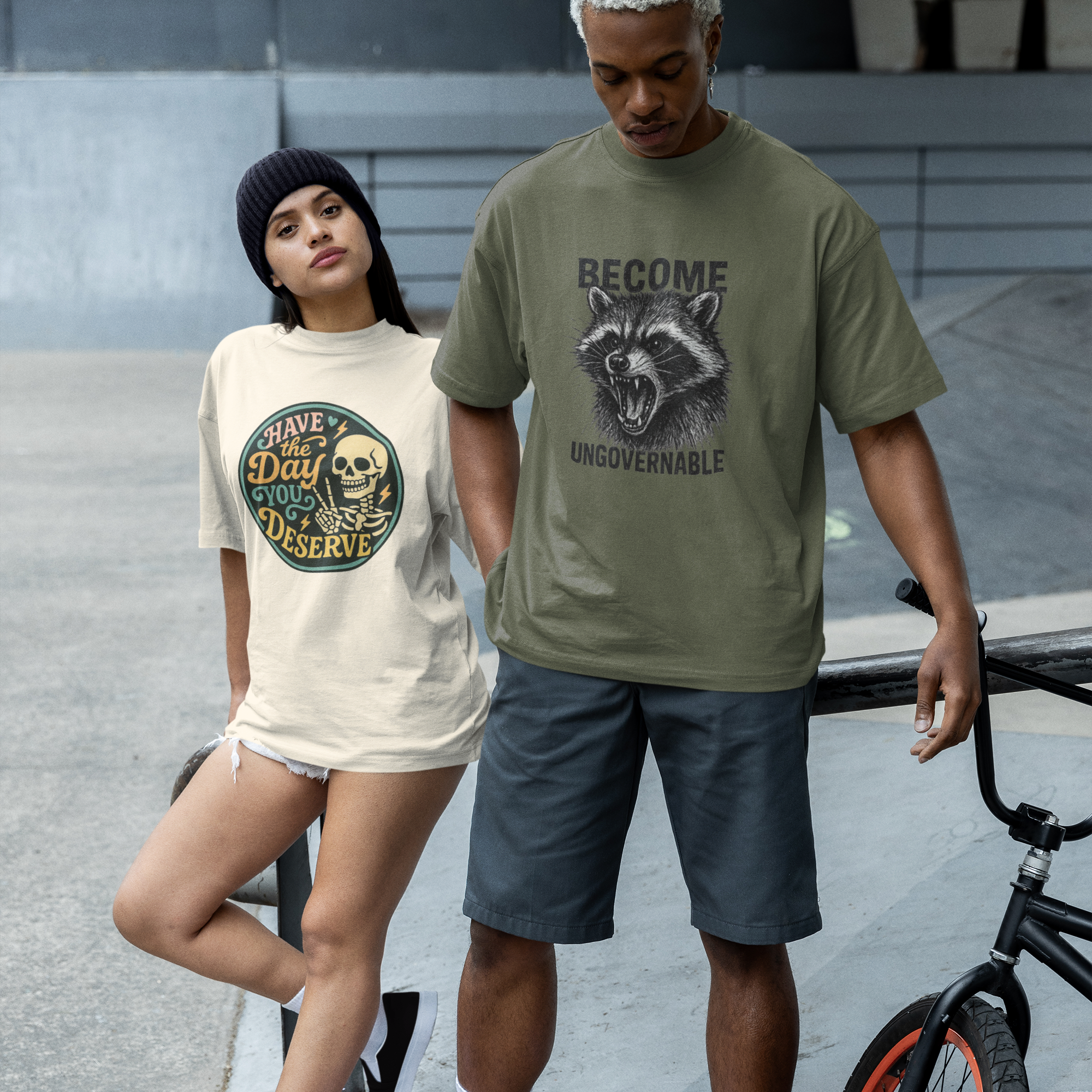Two people wearing Barely Functioning Co. graphic t-shirts, one in beige with skull design text 'Have the Day You Deserve' and one in olive green with raccoon design text 'Become Ungovernable,' standing outdoors near a BMX bike.